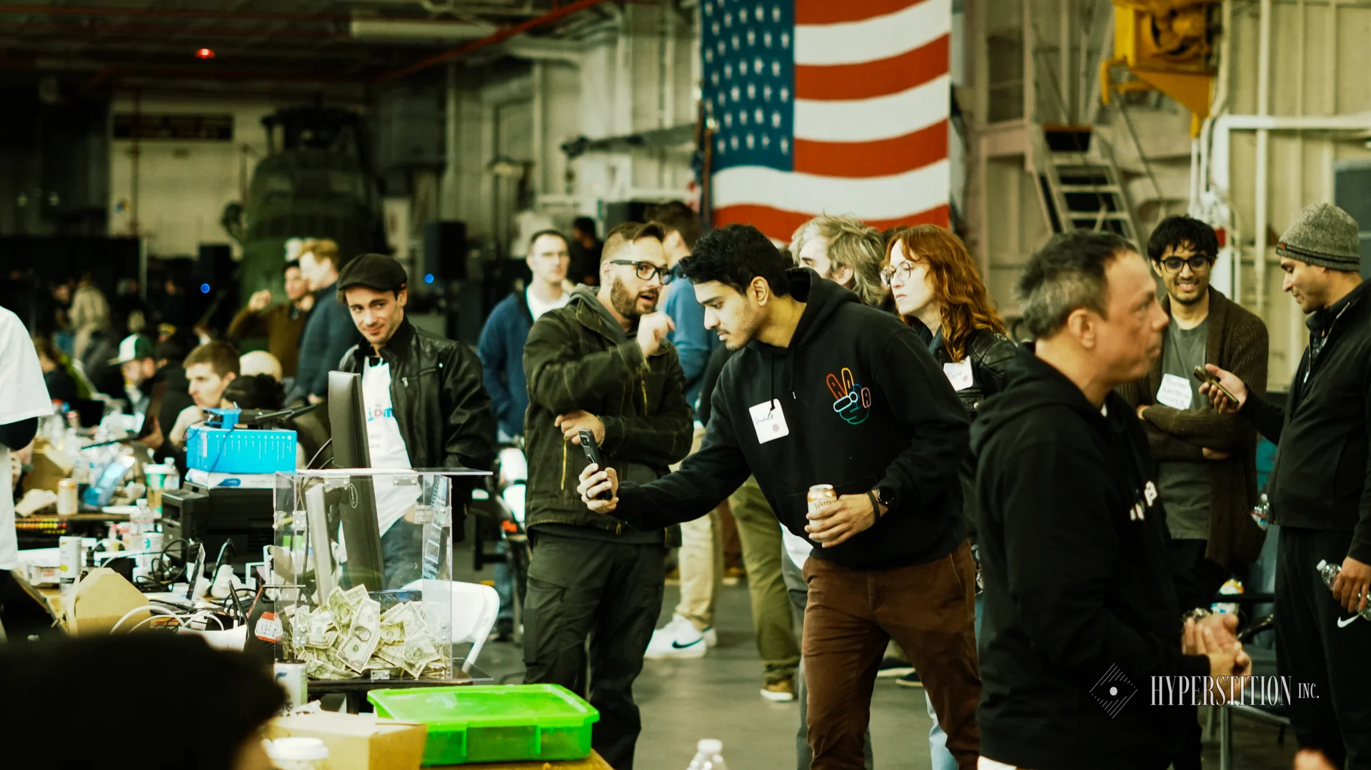 Builders gather around a hardware demonstration in the USS Hornet hangar during the 2024 hackathon